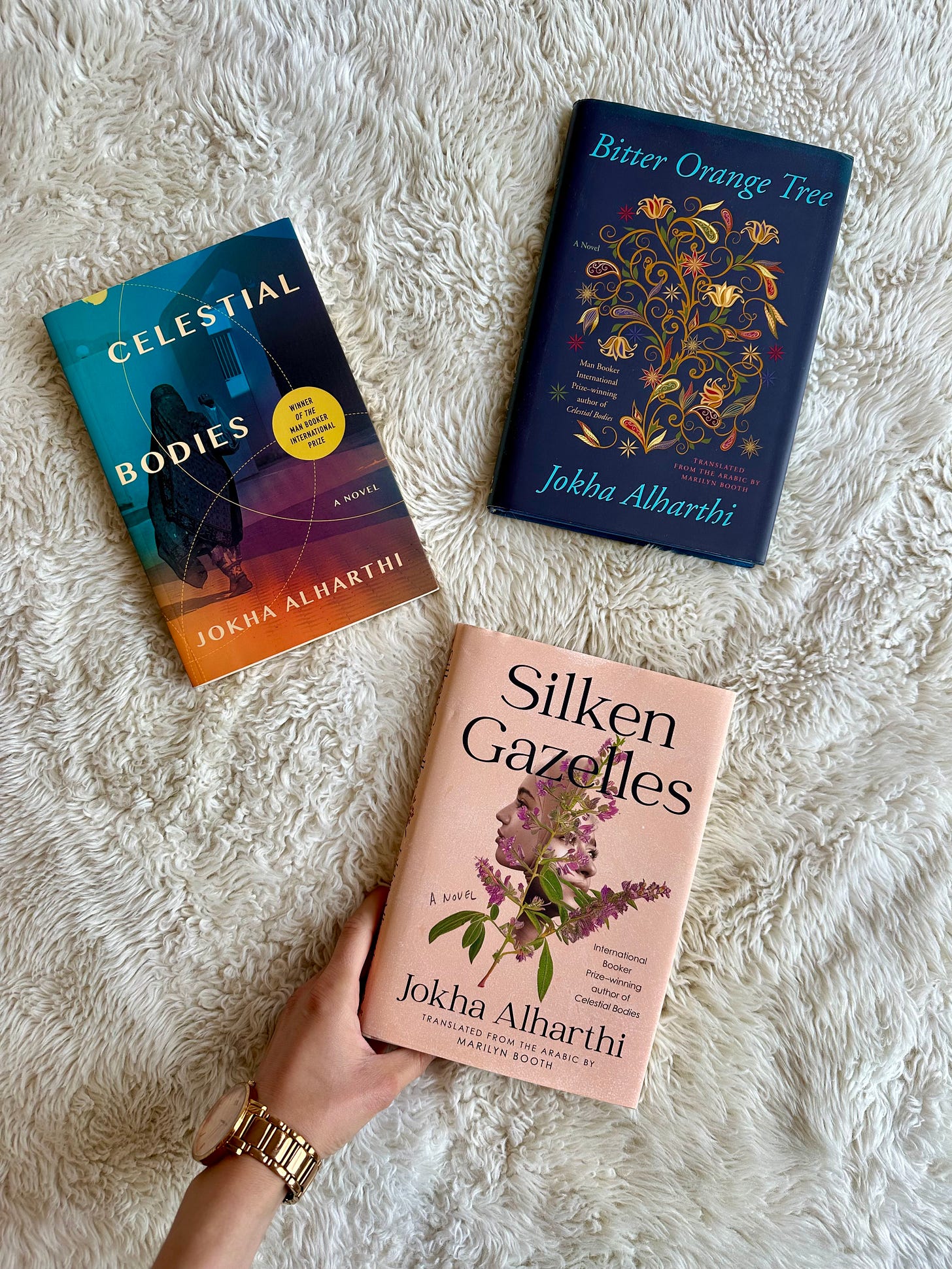 A white hand holds a copy of Silken Gazelles against a fluffy white blanket, next to Celestial Bodies and Bitter Orange Tree