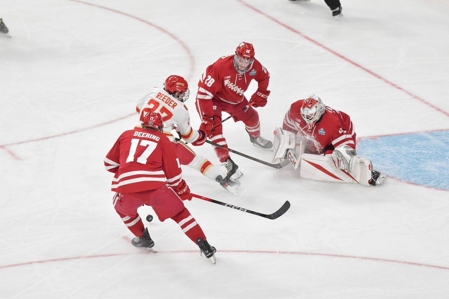 Hockey puck bounces in front of Wisconsin Badgers goaltender Daniel Hauser with Grady Deering and Aiden Dubinsky looking at it.