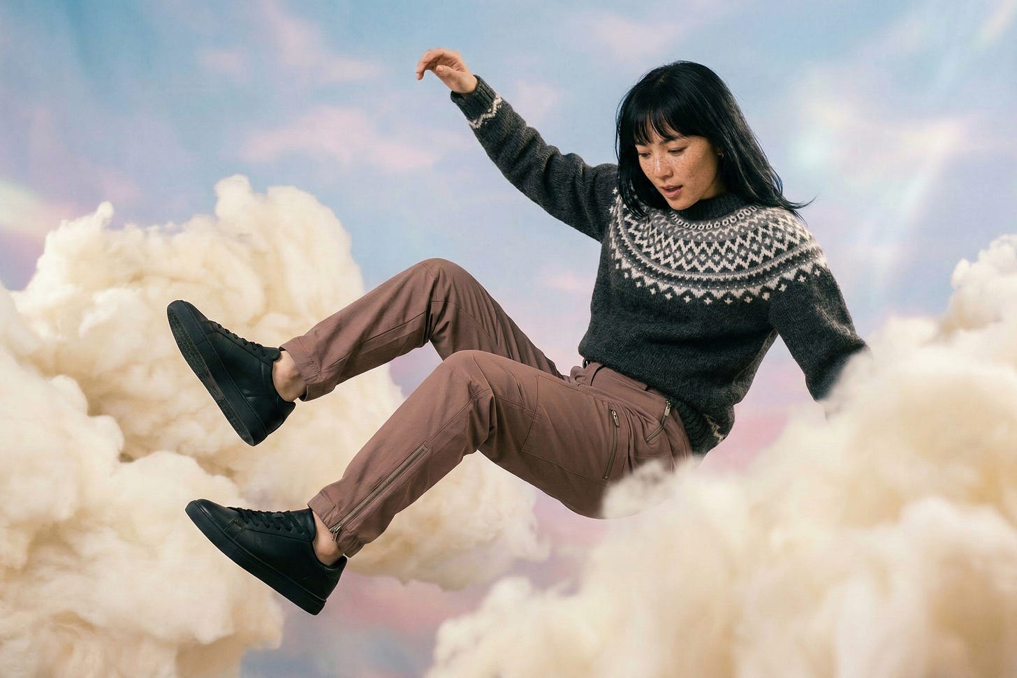 A high-fashion editorial portrait of a young woman mid-motion, captured in a dramatic pose as she falls through white and vanilla clouds floating in a pastel blue and pink dreamy sky at dawn. She wears an Icelandic jumper and muted clay-toned utility pants with subtle zipper details, paired with black sneakers.