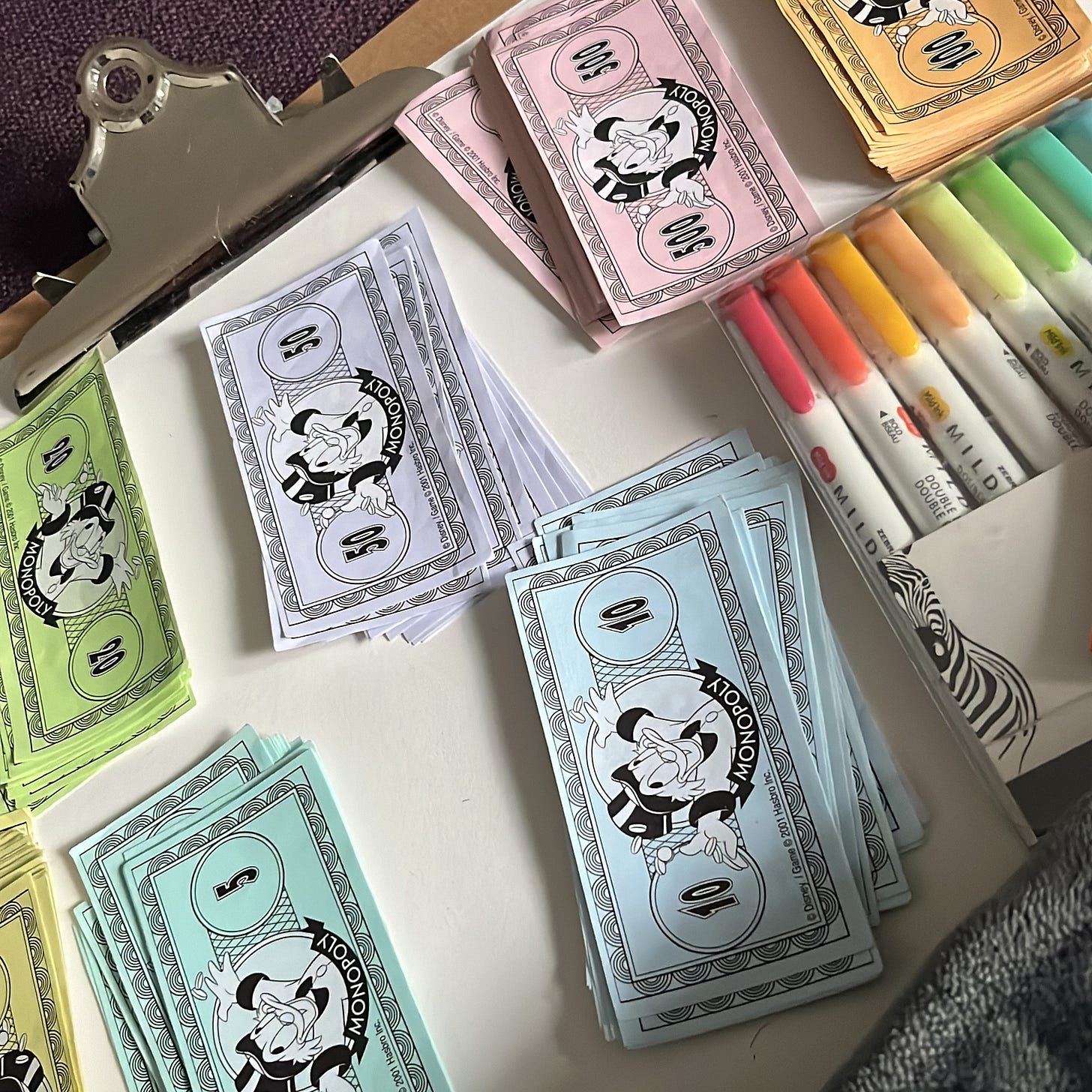 Piles of different coloured monopoly mony on top of a clipboard with some coloured pens