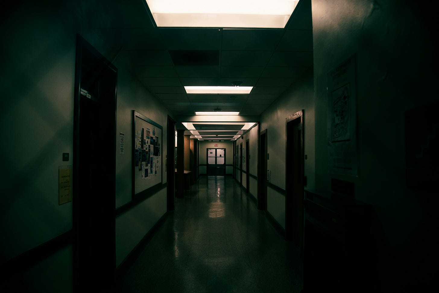 Dimly lit hallway of an institutional building, with closed doors lining both sides and fluorescent ceiling lights casting a harsh glow—evoking the atmosphere of a psychiatric hospital or forgotten ward.