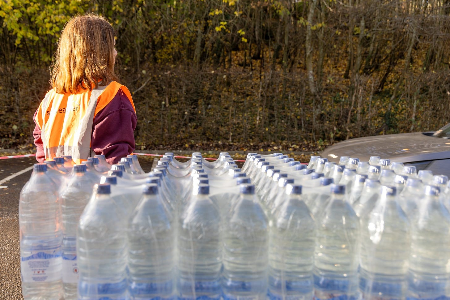 Person in orange safety vest stands near rows of bottled water stacked for distribution, in a car park with wooded area in front. A car is parked nearby.