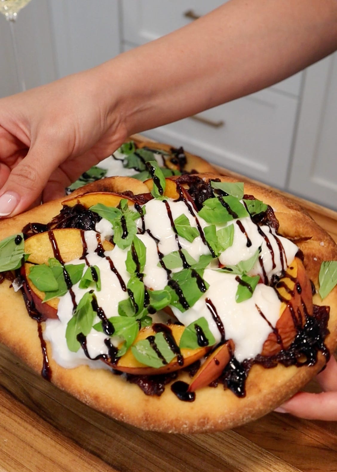 Close-up of a peach and burrata flatbread topped with caramelized red onions, fresh basil, and balsamic glaze being held over a wooden cutting board.