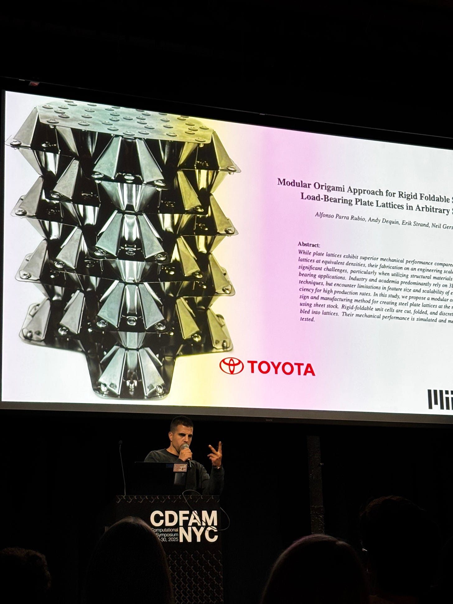 A presentation showing a modular origami approach for rigid foldable load-bearing plate lattices, featuring a metallic structure and speaker at an event.