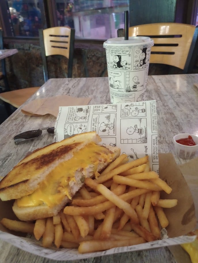 Soda cup with Peanuts strips, basket with sandiwch oozing cheese and a pile of fries, served on a paper covered in cartoons