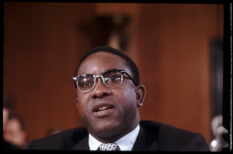 A seated man wearing a suit and glasses is photographed as he speaks. A seated man wearing a suit and glasses is photographed as he speaks.