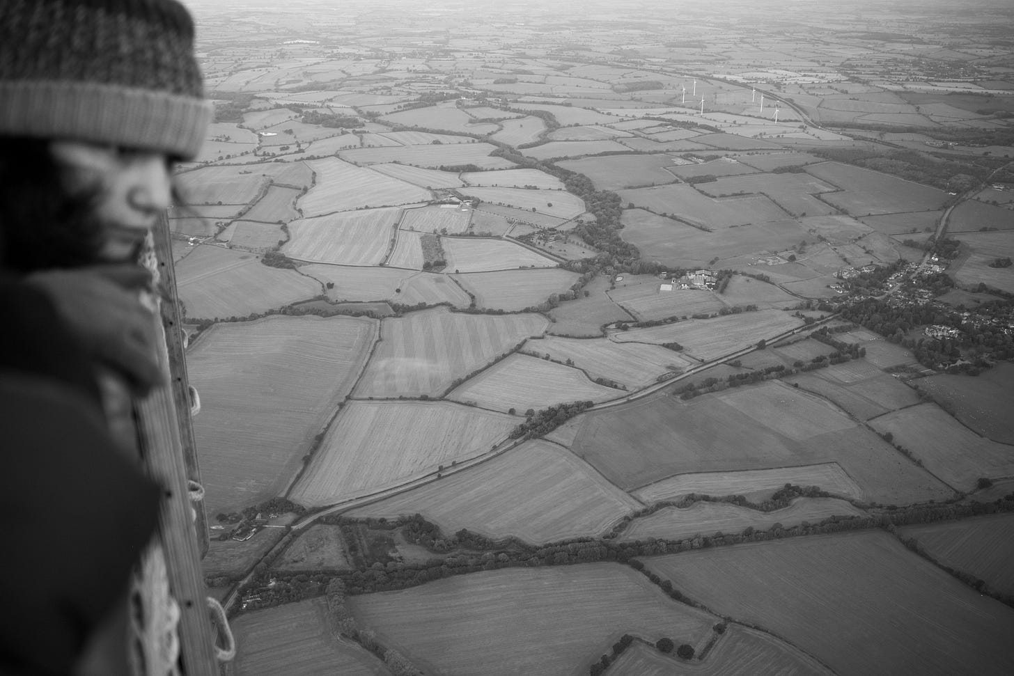 A young girl looks over the basket of a hot air balloon down across a tapestry of fields.