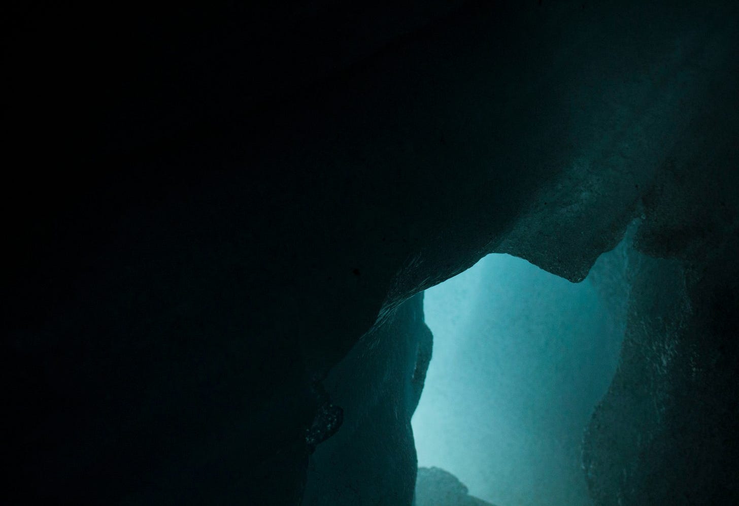 First-person view from inside a dark underwater cave, looking out through a narrow passageway towards a distant, hazy light.