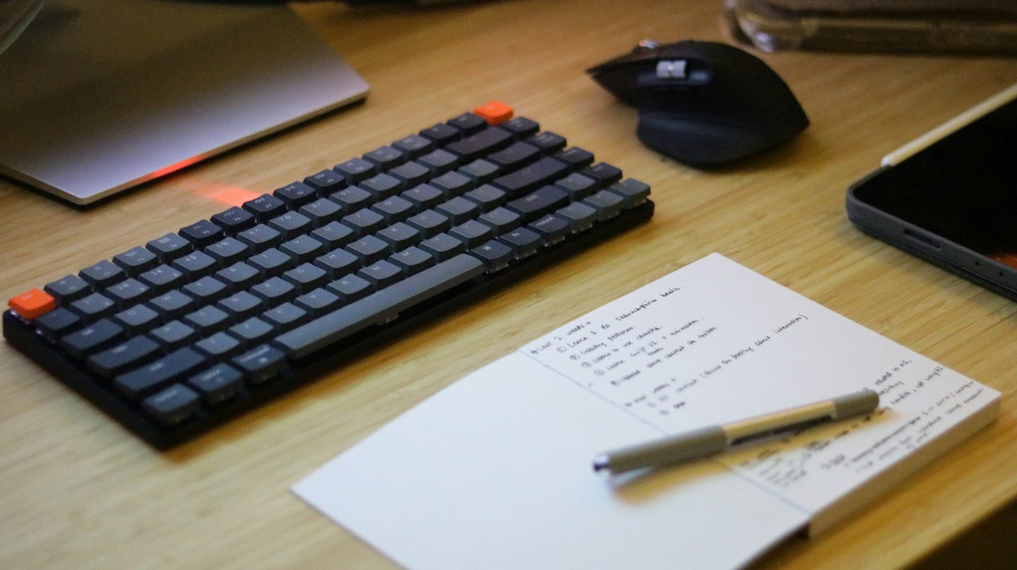 A light wooden desktop featuring a black keyboard and mouse, with an open notebook below the keyboard, sporting a pen and a series of handwritten notes. A light wooden desktop featuring a black keyboard and mouse, with an open notebook below the keyboard, sporting a pen and a series of handwritten notes.