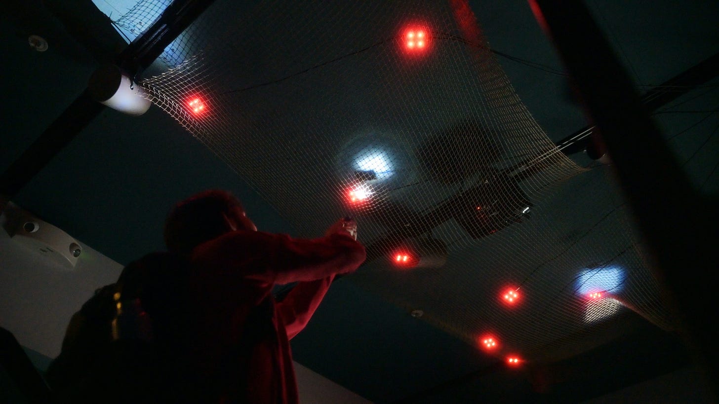 A silhouetted visitor points a flashlight up at a suspended white net glowing with red LED clusters in a dark gallery A silhouetted visitor points a flashlight up at a suspended white net glowing with red LED clusters in a dark gallery
