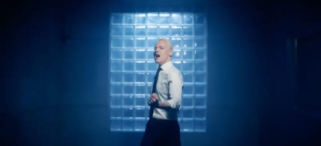 SHAMN, chemise blanche et cravate noire, blond platine, chante le poing fermé, seul devant une fenêtre même s'il est clairement éclairé de face SHAMN, chemise blanche et cravate noire, blond platine, chante le poing fermé, seul devant une fenêtre même s'il est clairement éclairé de face