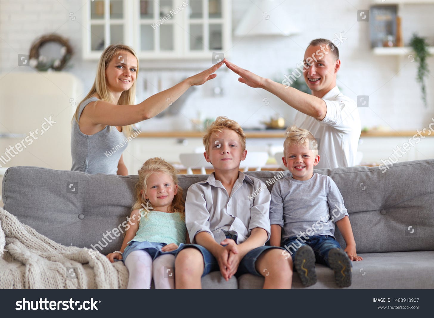 Happy full family with three kids sitting on sofa, mom and dad making roof figure with hands arms over heads. New building residential house purchase apartment and housing concept