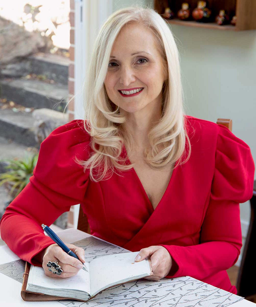 Woman with blonde shoulder-length wavy hair smiling at the camera. She is in a red longsleeved top sitting at her table with an open book and pen in hand, writing.