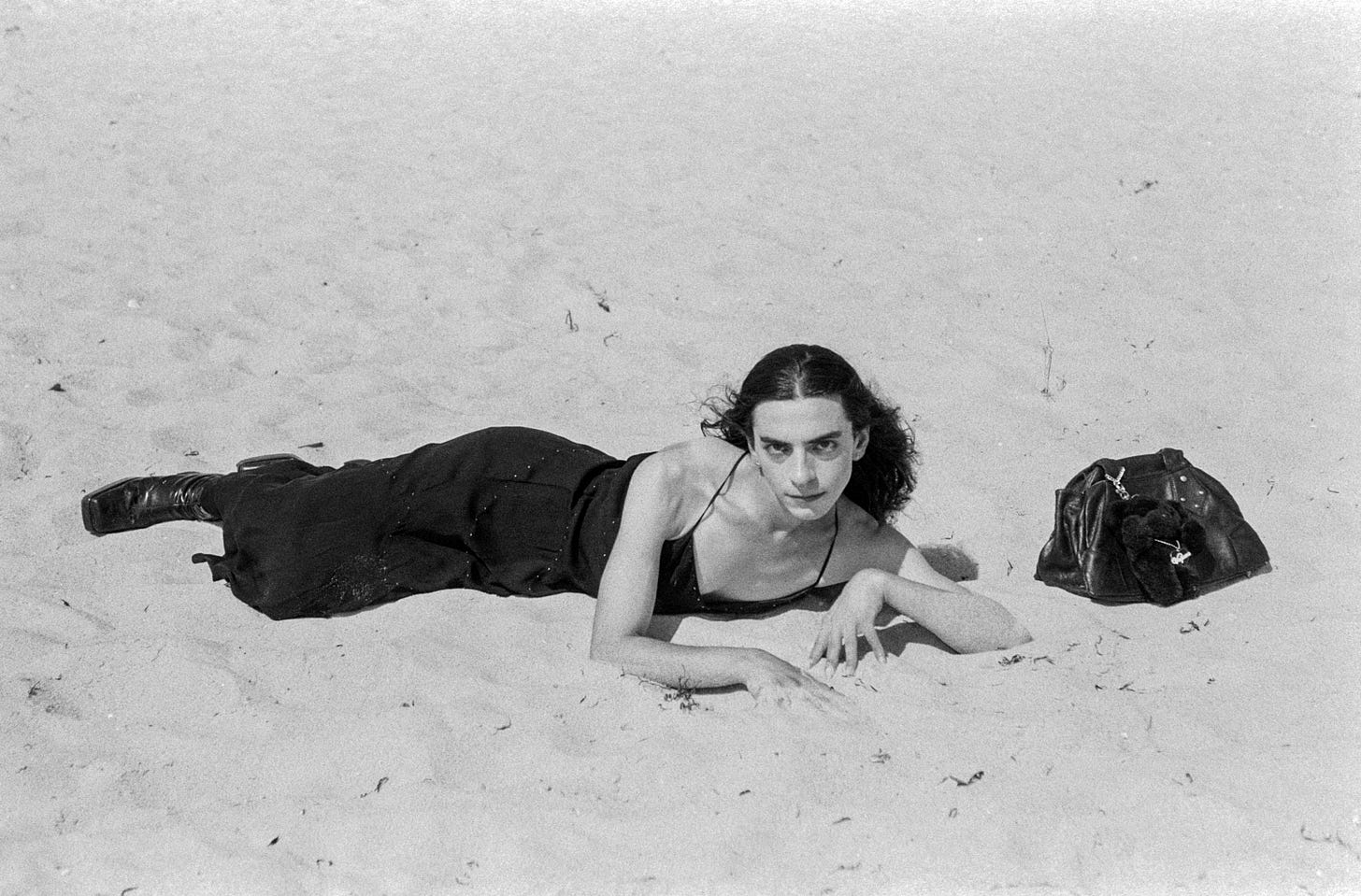 Artist lying in a black dress on sandy beach beside a bag, gazing directly at camera.