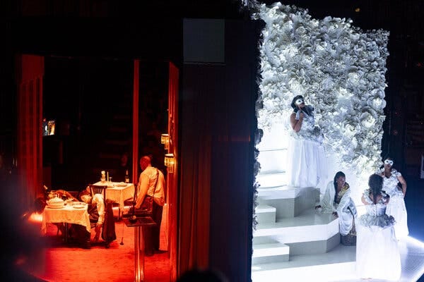 A stage divided in two with figures at a restaurant bathed in red light on the left, and figures dressed in white on a flower-draped staircase on the right.