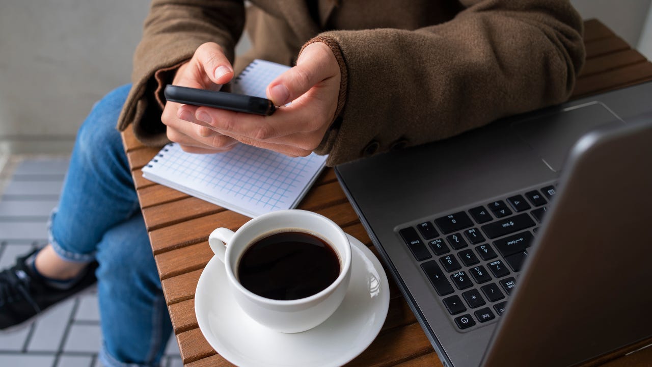 Close-up of a person holding a smartphone with a notebook, laptop, and a cup of black coffee on a wooden table.