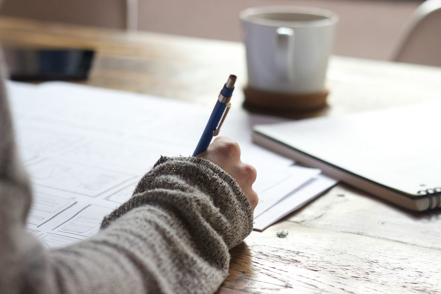 A woman's arm resting on a wooden table top, holding a mechanical pencil. Spread beneath the hand are a series of papers with writing and sketches. Beyond those rest a mug of coffee and a small spiral notebook.