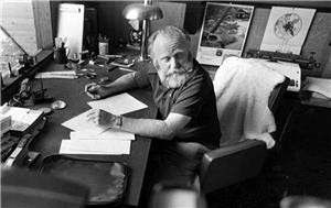 An older white man wearing a black t shirt, with a salt and pepper beard sits at his desk with a large book in front of him. There are pens, papers, and a lamp on his desk, and small posters on the wall in the background. 