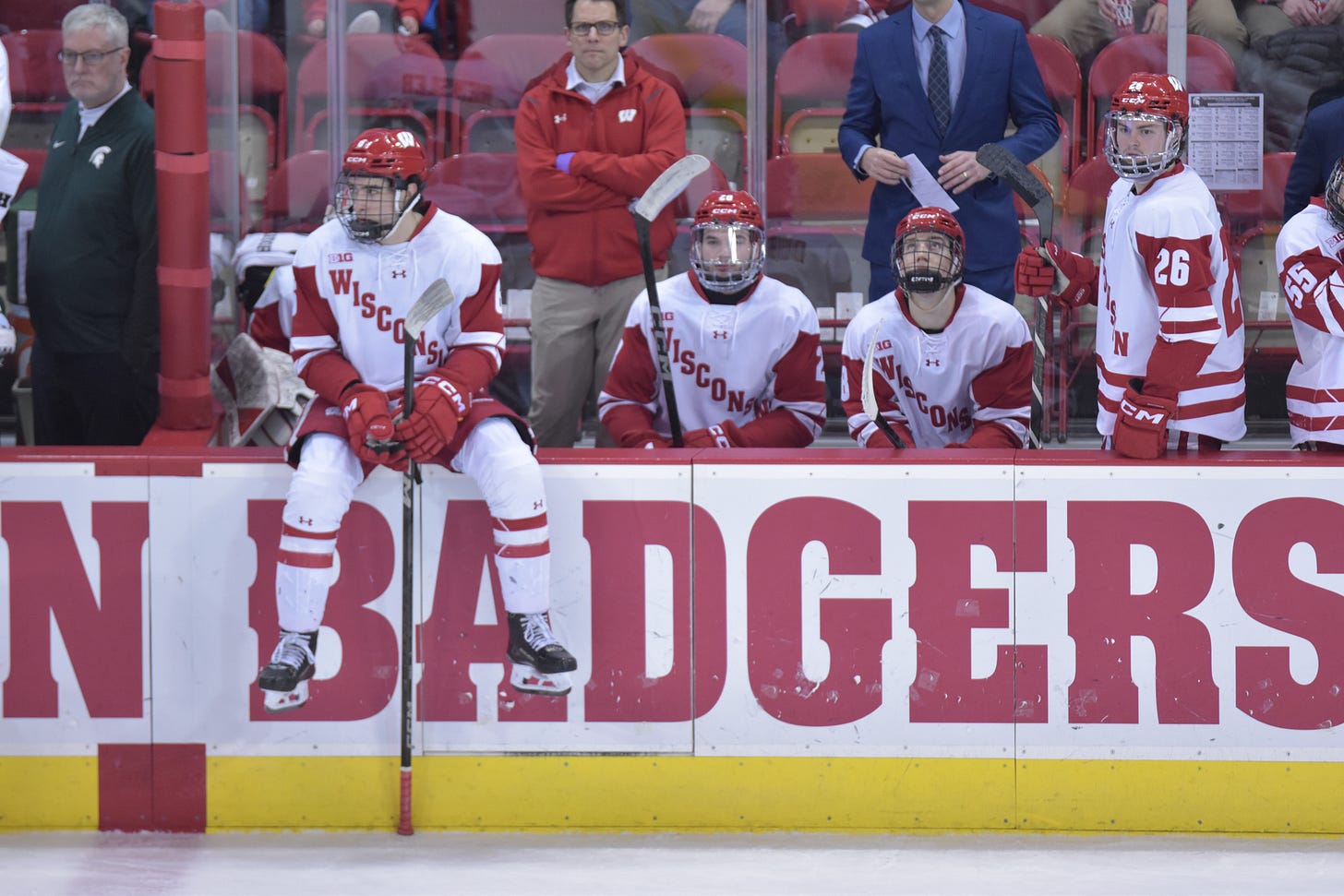 four wisconsin badgers men's hockey players are seen sitting on their team's bench with coaches and staff behind them