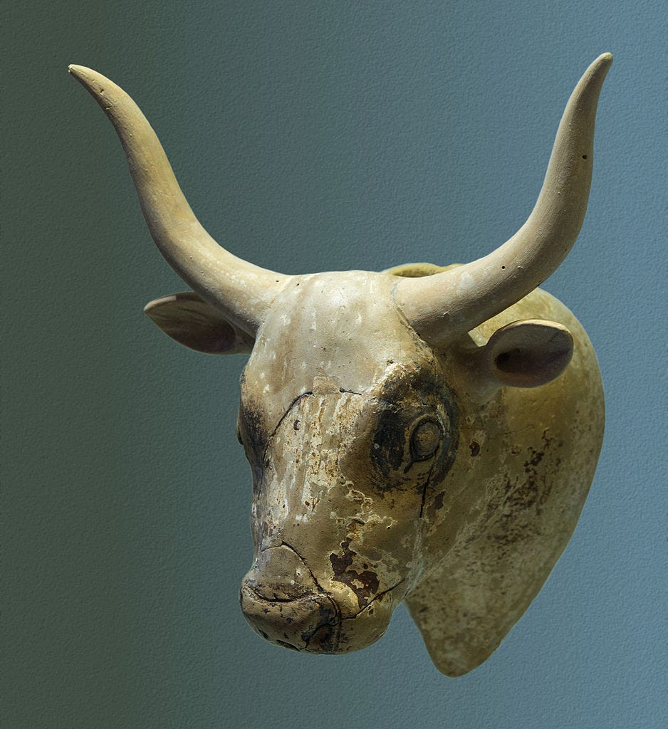 Ceramic horned cow head vessel. The vessel is made of tan ceramic with traces of white and brown paint remaining on it, showing that originally it was a white cow with brown spots on its neck and brown circles around its eyes.