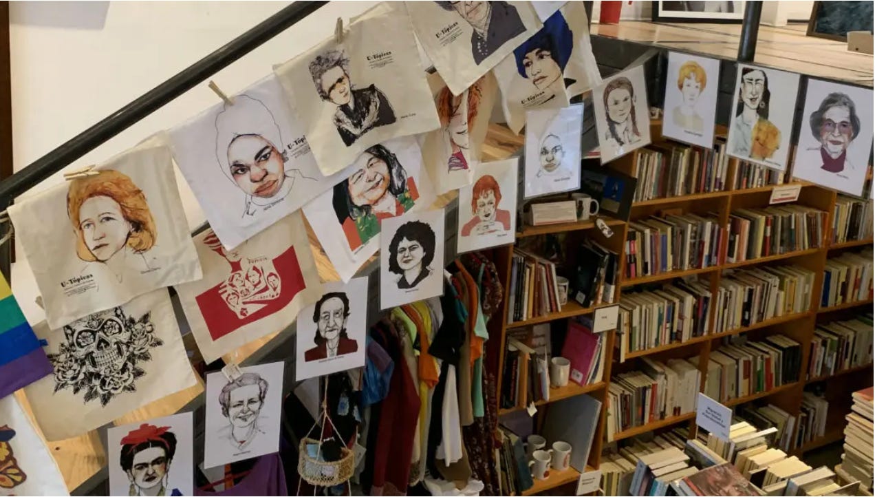 Un angolo della libreria U-Tópicas a Città del Messico. Sulla ringhiera di una scala sono appese diverse illustrazioni che ritraggono artiste e attiviste. Si riconoscono Frida Khalo, Mercedes Sosa, Angela Davis. Sullo sfondo, scaffali pieni di libri.