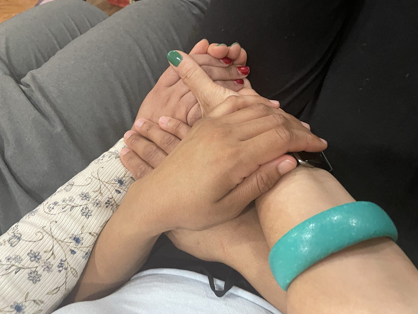 three hands in various shades of light to medium brown hold each other, two have nail polish- one cherry red, one teal green and silver, on a backdrop of grey pants/ a white waffle knit fabric over an arm with grey flowers on it.