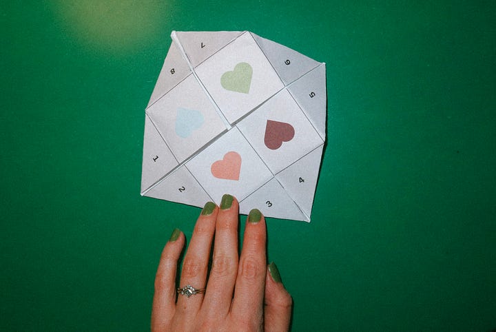 Hands holding a paper fortune teller decorated with pastel hearts; the fortune teller laid out against a green background.