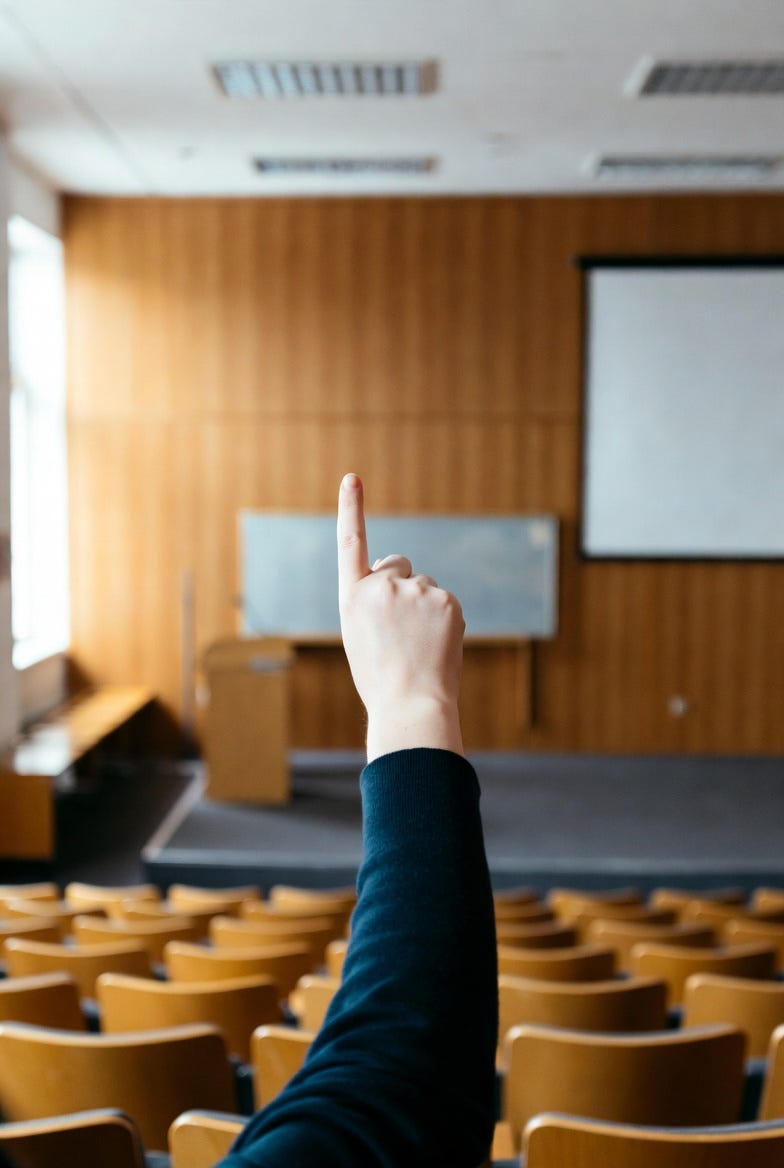 A single hand raised in the back of an empty lecture hall, fingertip pointing up toward the stage and projection screen.