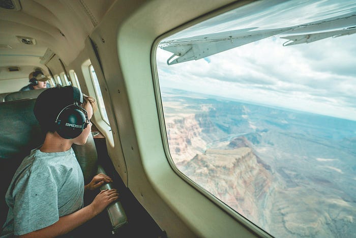 A boy staring out the window with headphones on overlooking the mountains while on a plane.