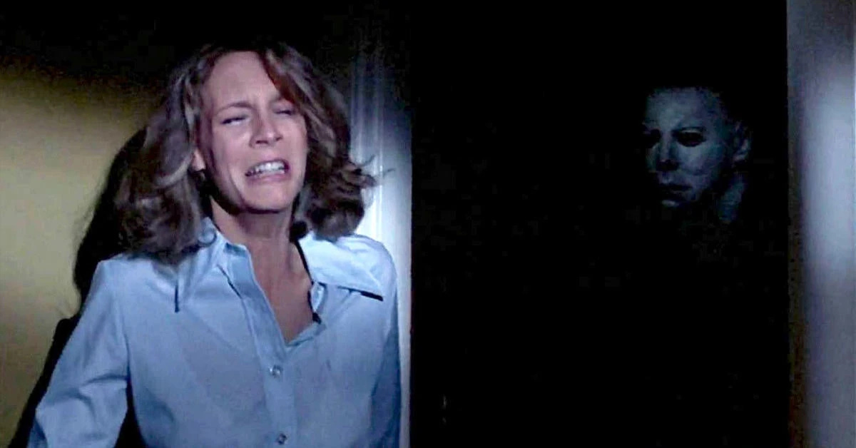 Laurie Strode in Halloween; she's wearing a blue shirt with the top button undone and is crying as she stands against the wall; Michael with his mask is in shadow in the doorway Laurie Strode in Halloween; she's wearing a blue shirt with the top button undone and is crying as she stands against the wall; Michael with his mask is in shadow in the doorway