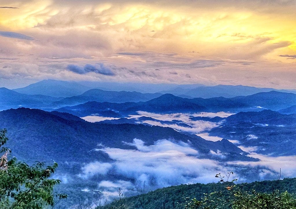 A view of hills from above the clouds, with sunset burning in the top half of the picture above the blue-grey hills and valleys in the bottom half.