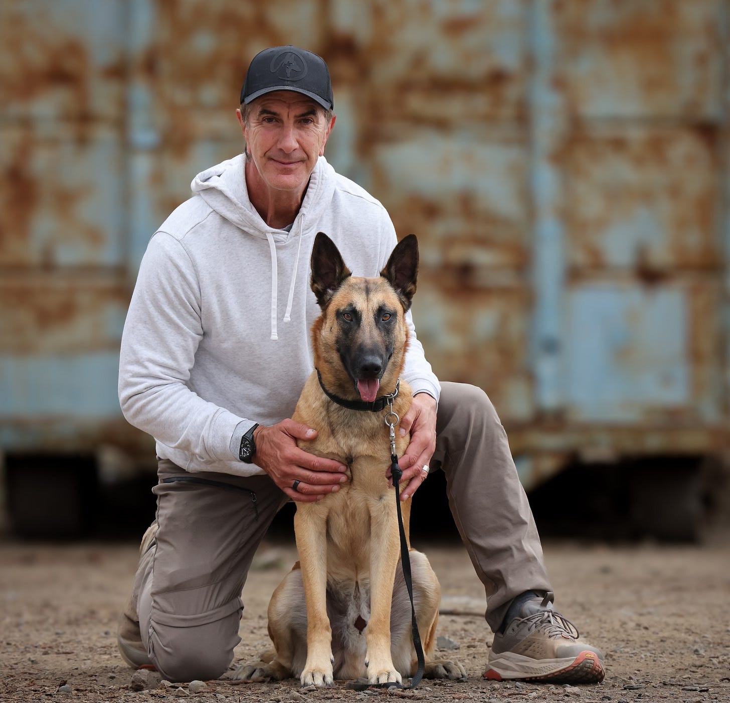 Dog trainer Robert Cabral kneels beside a tan Belgian Malinois-type dog on leash outdoors.