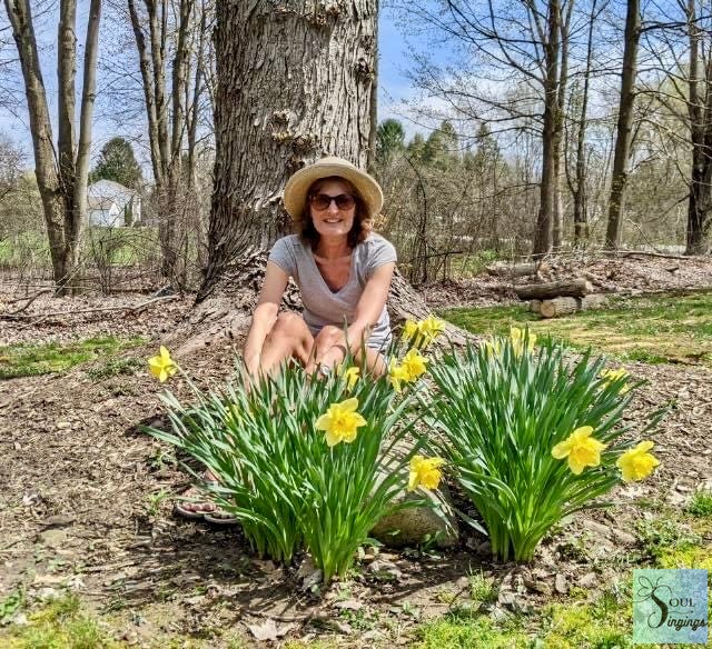 60 Things I've Learned at 60: author in a hat crouched behind bunches of yellow daffodils