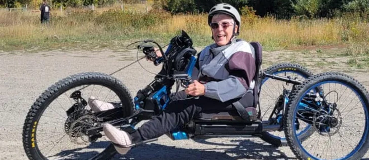 A smiling person wearing a helmet and sunglasses rides a blue off-road handcycle on a sunny day. The individual is on a gravel path surrounded by grassy fields and trees, dressed in a light jacket and dark pants, appearing to enjoy the outdoor adventure.