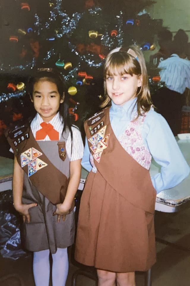 Two elementary age girls stand in front of a large decorated Christmas tree wearing Girl Scouts uniforms.