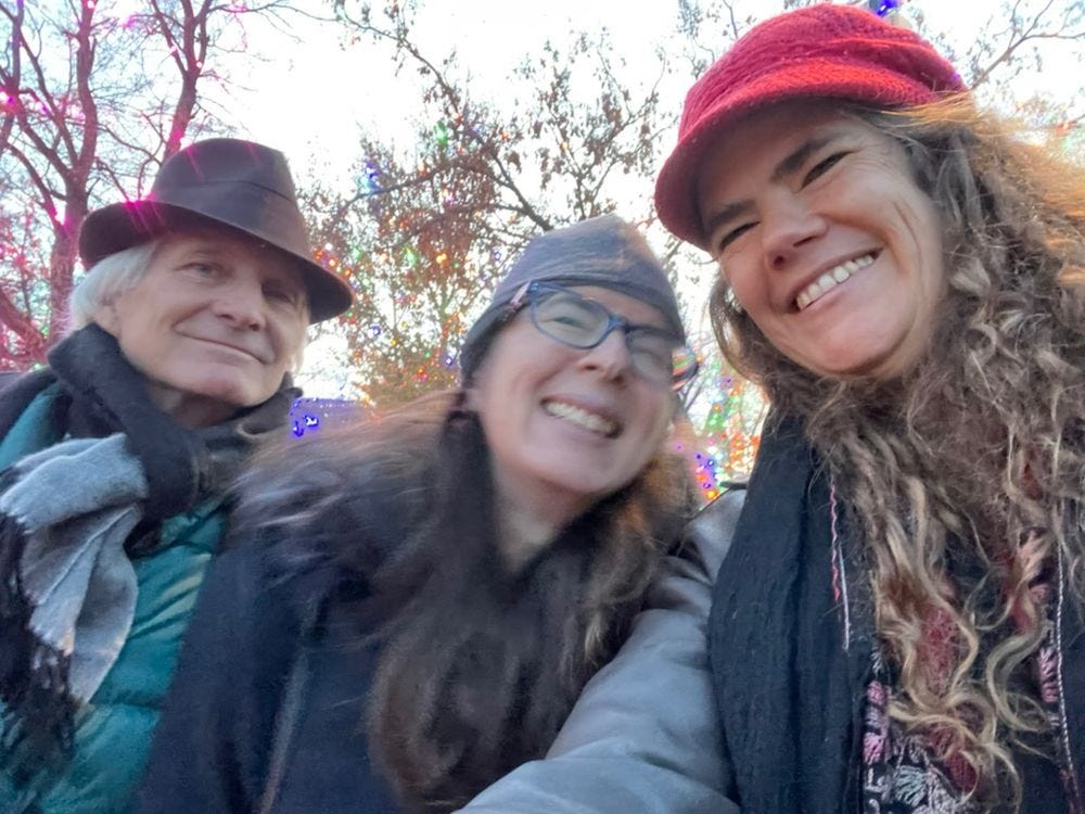 A white man (John Kadlecek) in a natty brown hat and a black and gray scarf, a white woman (Beth Mazur) with long brown hair in a gray cap, slightly blurry from movement, and a white woman (Julie Rehmeyer) with curly brown hair and a red hat, all smiling, with trees in the background covered with Christmas lights