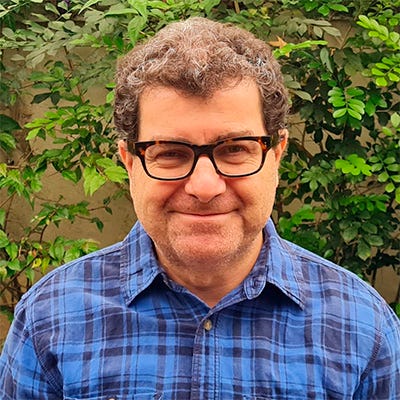 Homem branco, sorridente, com óculos de armação preta. Ele usa uma camisa quadriculada azul. O ambiente é externo e, ao fundo, observam-se algumas plantas.