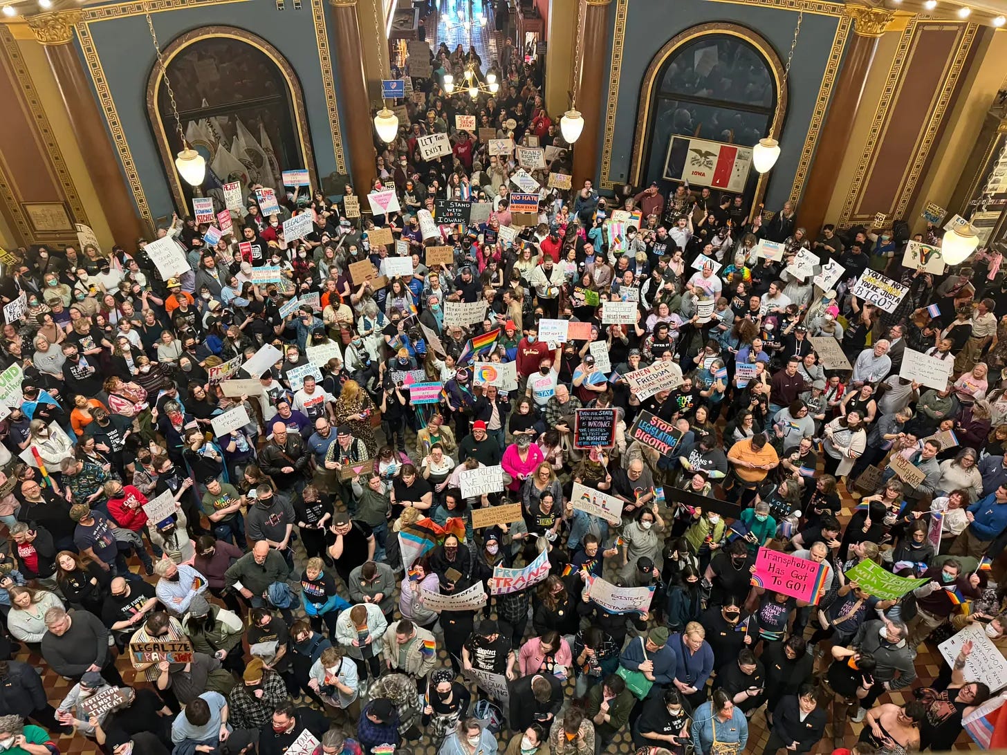 Alt text: Overhead photograph capturing a large, dense crowd of hundreds of protesters filling the multiple levels of the Iowa State Capitol rotunda. Diverse people hold signs protesting SF 418 and advocating for transgender civil rights. The image conveys the significant scale of public opposition to the legislation. Alt text: Overhead photograph capturing a large, dense crowd of hundreds of protesters filling the multiple levels of the Iowa State Capitol rotunda. Diverse people hold signs protesting SF 418 and advocating for transgender civil rights. The image conveys the significant scale of public opposition to the legislation.