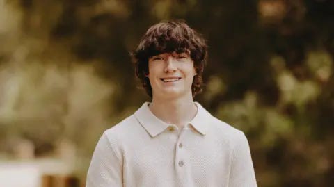 The Raine Family A photo of Adam Raine. He has long, brown shaggy hair that is wavy. He is seen smiling, wearing a knit collared shirt with three buttons at the top. Behind him is a blurred background of foliage. 