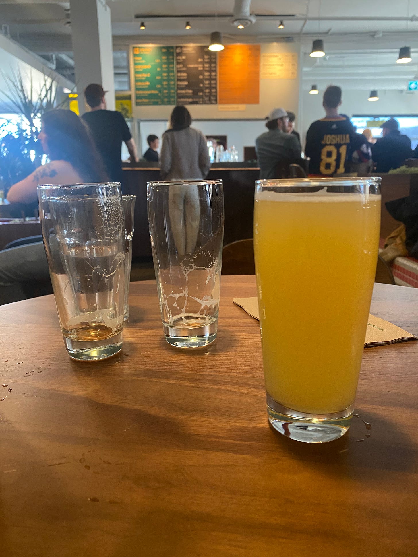 Brewery interior: foreground shows a round wood table with a 12oz glass of IPA and and a couple of empty glasses behind it. In the background is the bar, with people standing in front of it, and the colour-coded tap list for each brewer in behind (green, black, and orange).