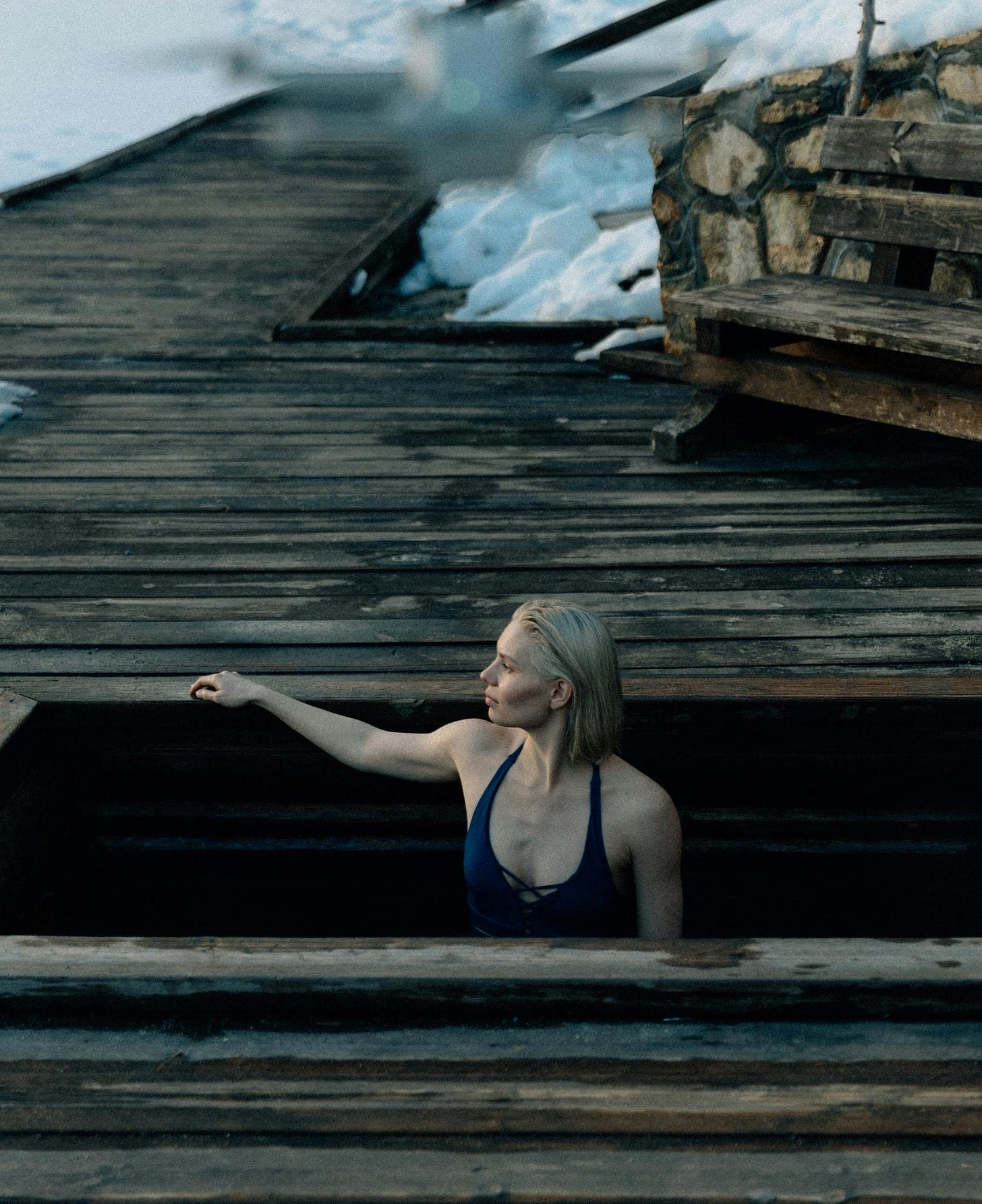 A woman in a blue swimsuit, with short blonde hair, sits partially submerged in a cold plunge pool by a lakeside. Wooden decking and snowy mountains in the background emphasize the cold outdoor environment. A woman in a blue swimsuit, with short blonde hair, sits partially submerged in a cold plunge pool by a lakeside. Wooden decking and snowy mountains in the background emphasize the cold outdoor environment.