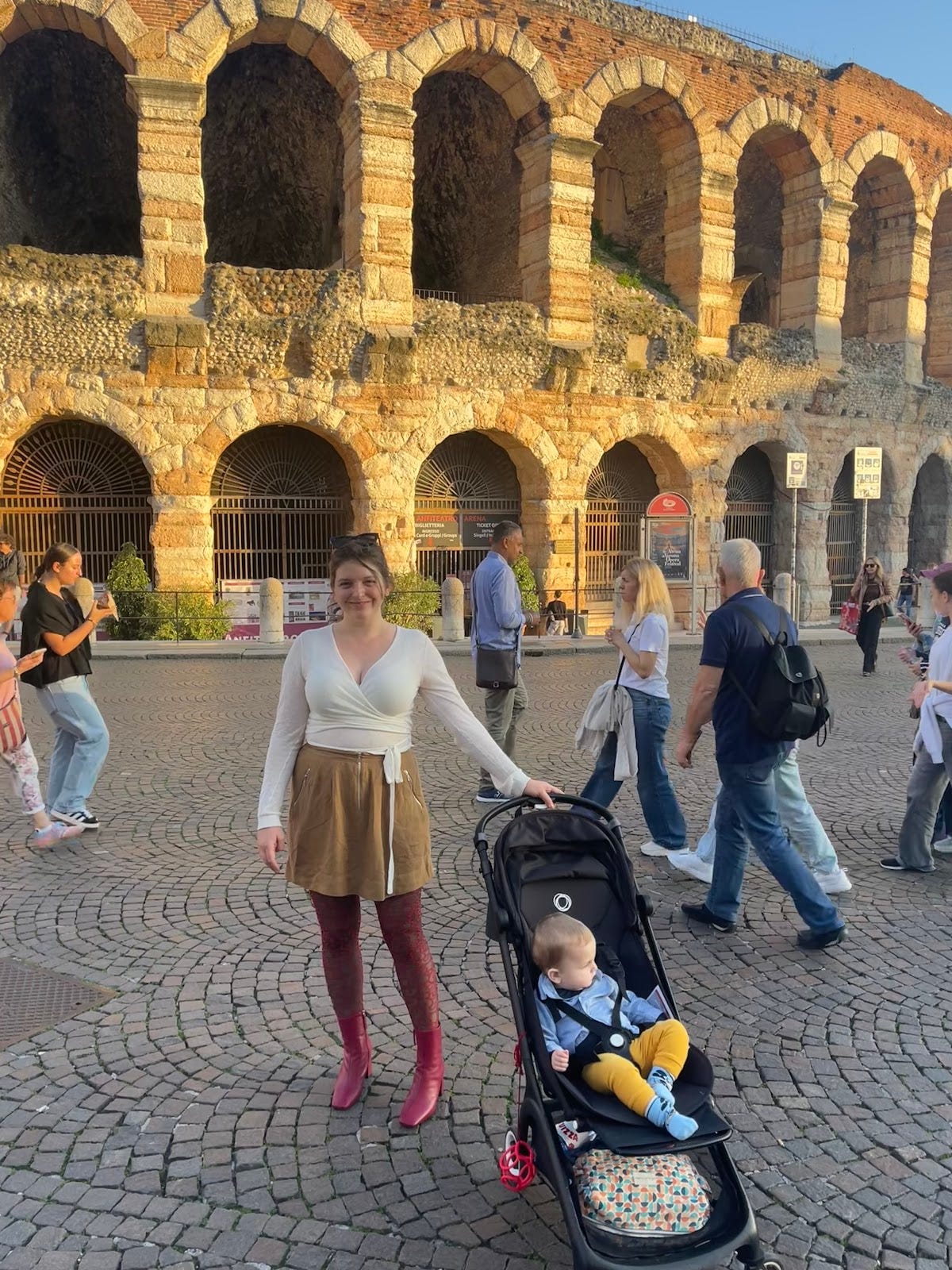 The author standing in front of the arena in Verona, her baby in a buggy, in the evening sun. She is wearing awesome pink boots. 