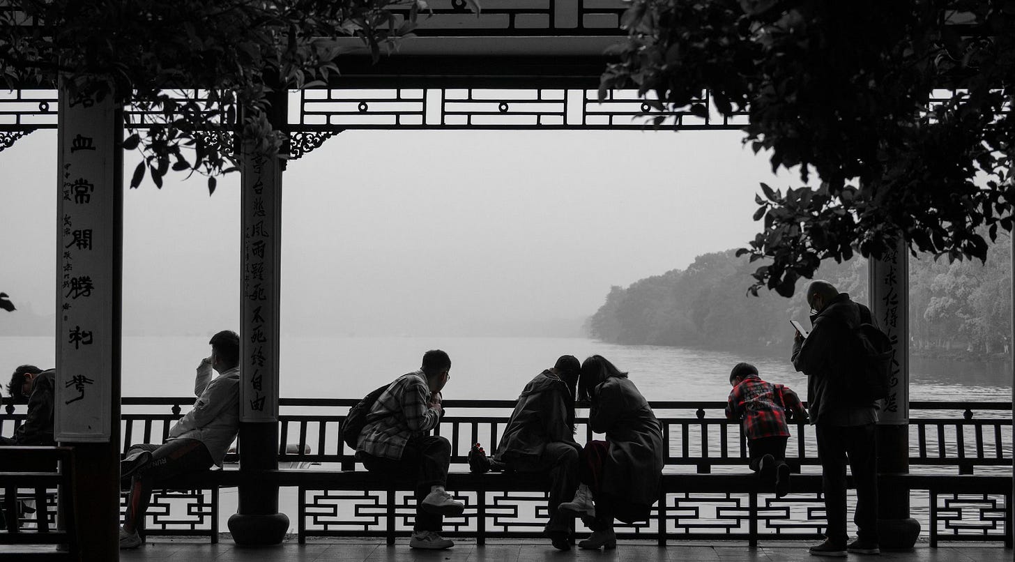 Fotografia, a maior parte dela em branco e preto, de pessoas em uma bela varanda com arabescos chineses com vista para um rio – no entanto, nota-se o vermelho da camisa de um menino.