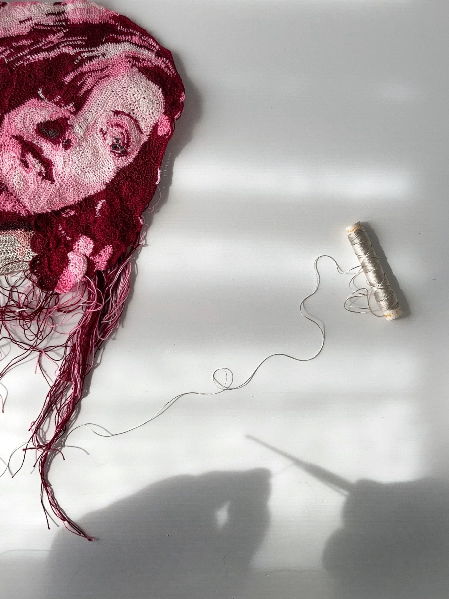 Shadow of two hands holding a crochet hook on a white surface where lays flat a crochet portrait of Greta Garbo and a spool of thread