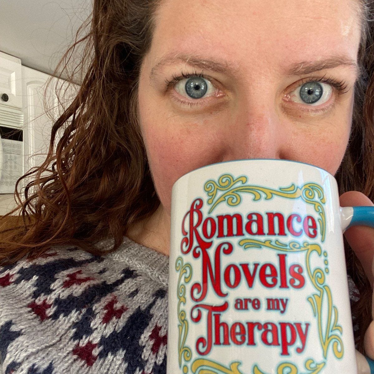 A selfie of a woman with red hair and blue eyes sipping a mug that says "Romance Novels are My Therapy"