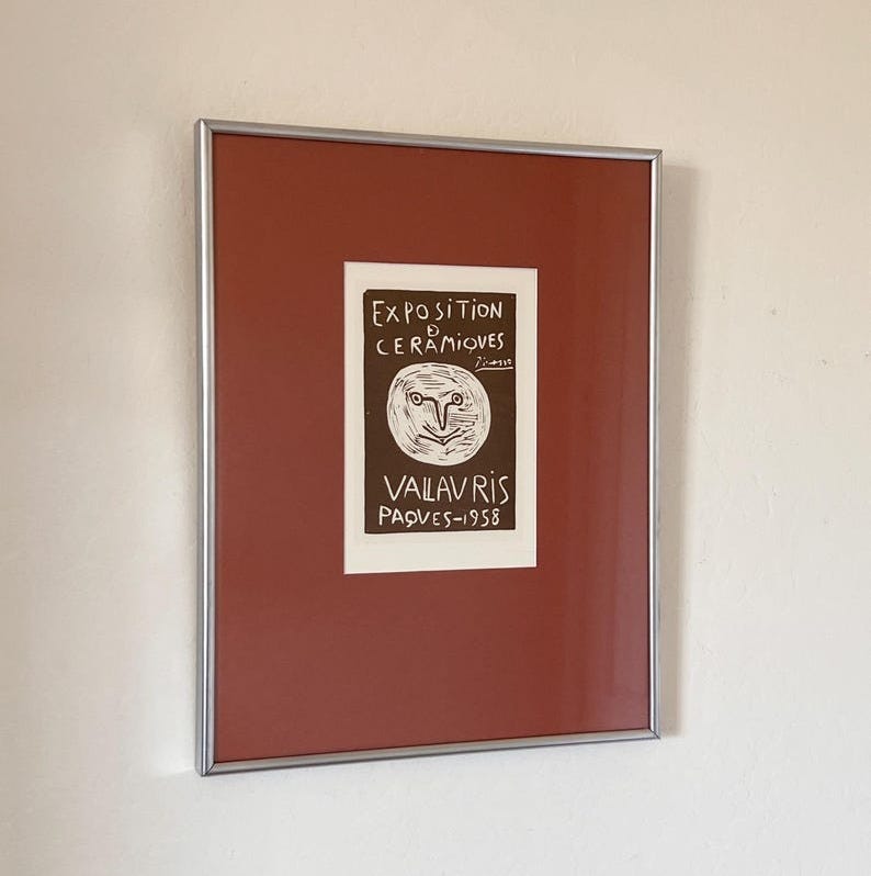 May include: A framed poster with a brown background and white text. The text reads 'Exposition de Céramiques', 'Picasso', 'Vallauris', 'Pâques-1958'. There is a drawing of a face with a large nose and eyes.