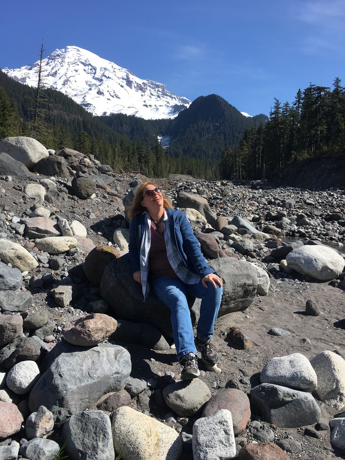 A woman sitting on a rock in a dry river bed looks up at the sky, a snow-covered mountain rises against blue sky behind her.