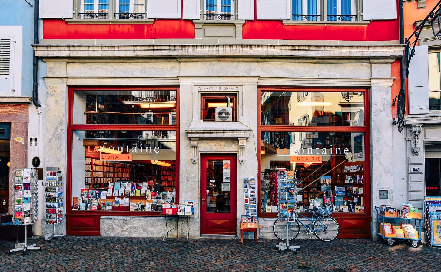 facade of a bookstore with large plate-glass windows and a bright red door. there's a blue bicycle parked out front and racks of colorful postcards.