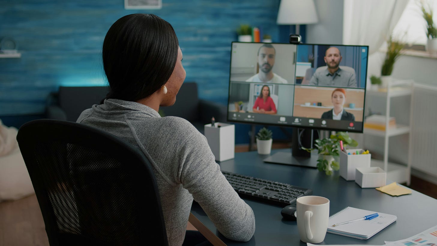 A non-white person sits at a desk and looks at a screen displaying four people in a virtual meeting. Her back is to the camera and she has long black hair, is wearing a gray long-sleeved shirt, and has a mug of tea in front of her. A non-white person sits at a desk and looks at a screen displaying four people in a virtual meeting. Her back is to the camera and she has long black hair, is wearing a gray long-sleeved shirt, and has a mug of tea in front of her.