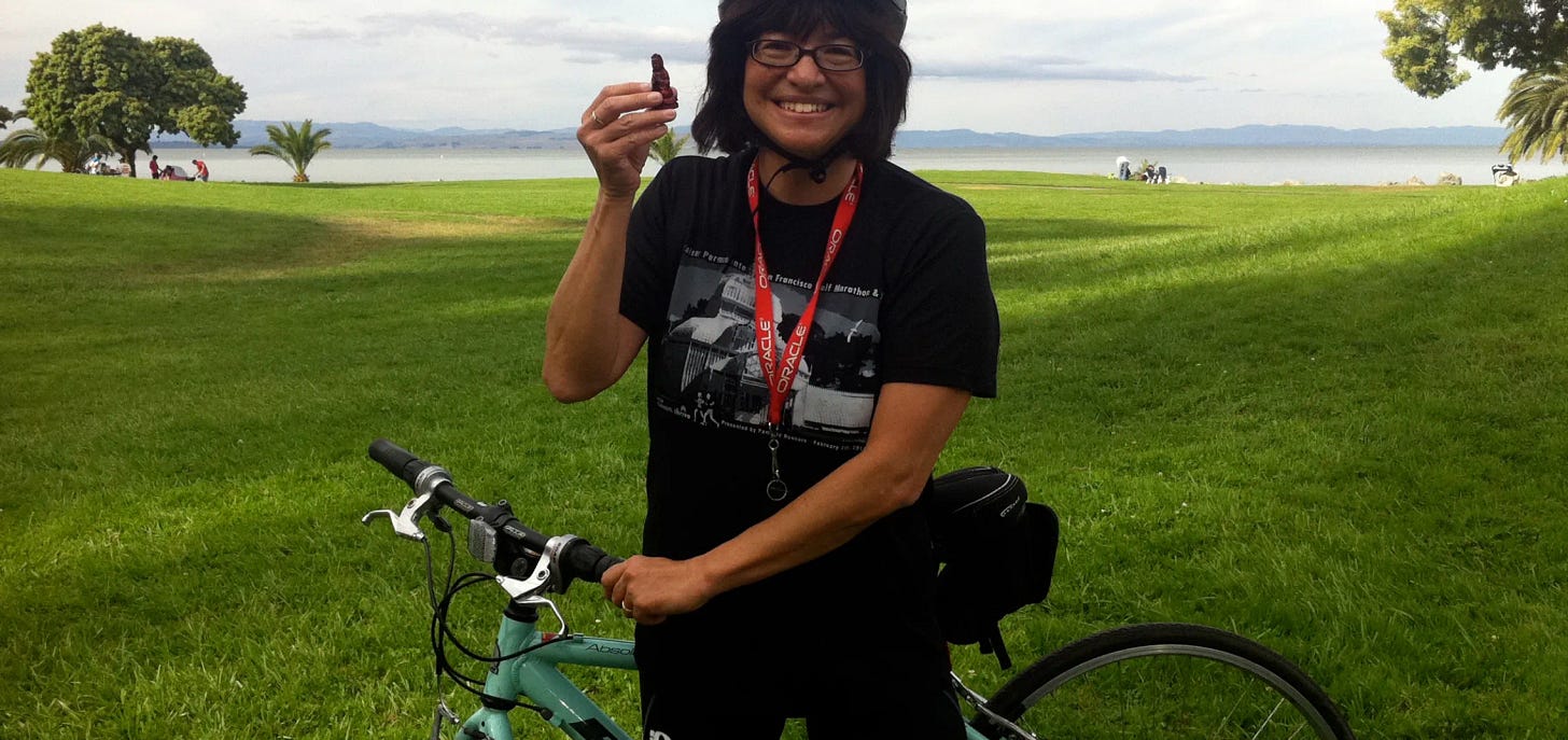 A woman in a bike helmet standing with her bicycle and smiling on a grassy field with water in the background